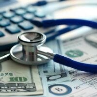 A close-up of medical expenses concept with a stethoscope, calculator, and US dollar bills on a medical billing statement, depicting the financial aspect of healthcare. Close-up view of a stethoscope resting on a surface with US dollar bills and a calculator.