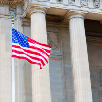 The image shows the American flag waving in front of a neoclassical building with large stone columns, conveying patriotism and historical significance.