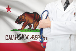 Doctor in a white coat holding a stethoscope, standing in front of the California state flag featuring a bear and red star, conveying healthcare professionalism.