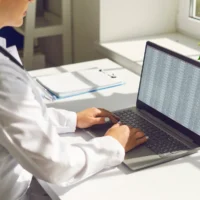 Doctor in office with laptop showing error. Healthcare computer shows corrupted code, malware or cybersecurity issue while professional works at desk. Medical data security and breach concept.