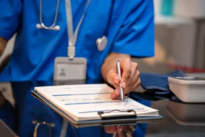 Close-up of healthcare worker in blue scrubs filling out medical forms on clipboard at desk, with stethoscope and badge visible, clinical office setting.