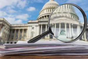 Close-up of a magnifying glass over a stack of official documents on a wooden desk, symbolizing scrutiny and transparency in legal or investigative processes.
