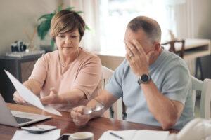 Older couple reviewing household bills and finances at a table with a laptop and calculator; the man covers his face in stress.